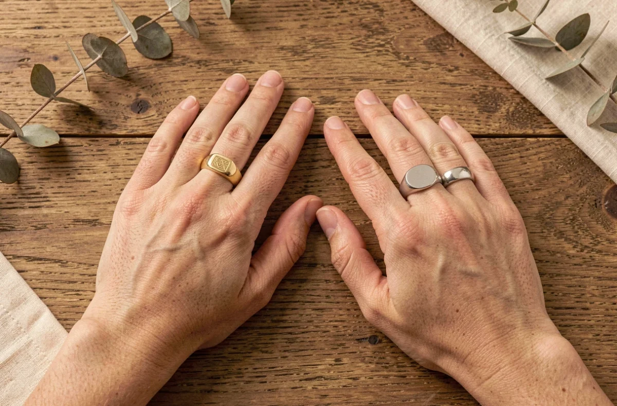 Comparaison de deux chevalières minimalistes portées côte à côte, l'une en or et l'autre en argent, sur une table en bois brut